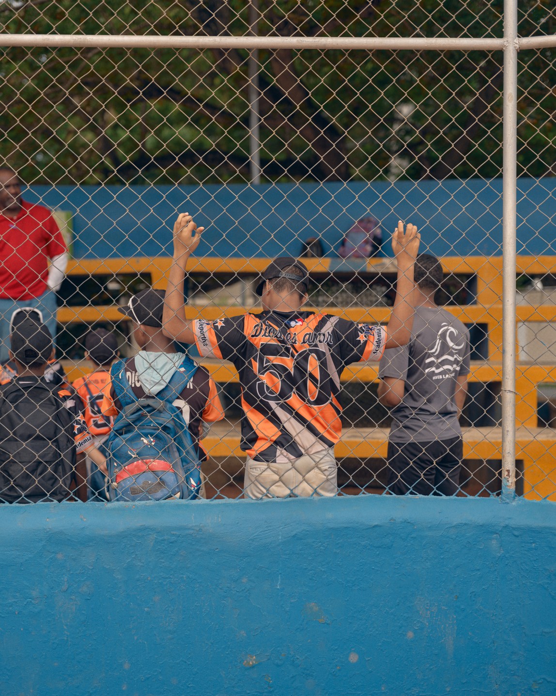 A young baseball player in an orange-and-black uniform grasps the chain-link fence behind him, leaning into it with his back. Other kids stand near him in front of orange bleachers.