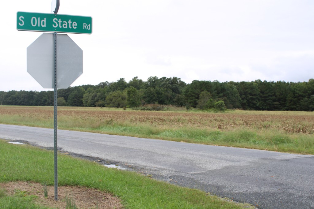 A roadway and open field near a street sign that reads S. Old State Street.