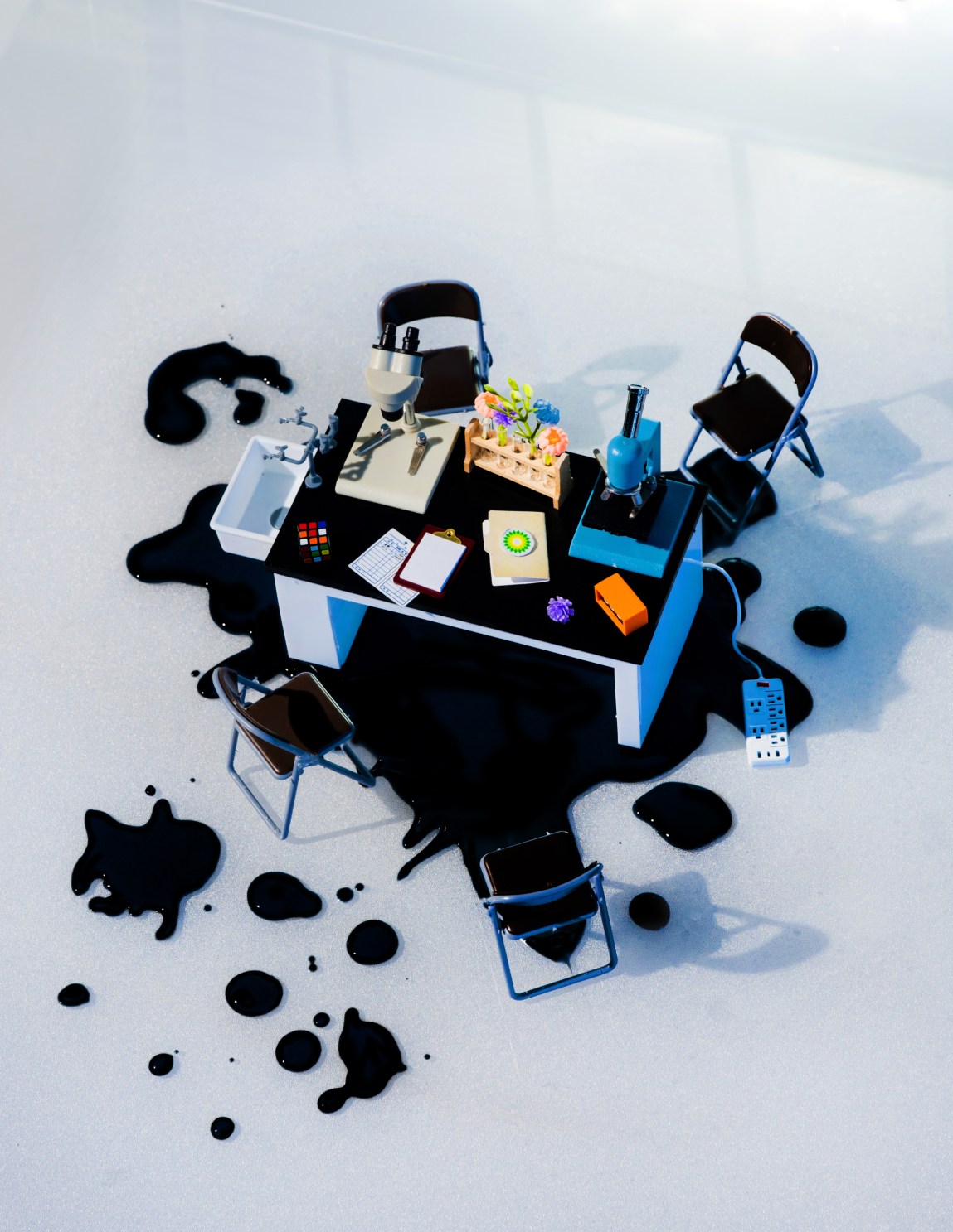 A miniature desk with flowers, microscopes, folders and office supplies sits on a pool of splattered black oil.