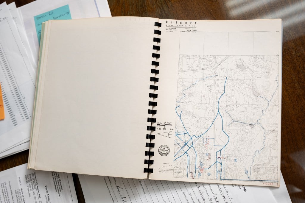 A spiral bound book, opened to a page showing a topographic map with blue lines, laying on a desk covered with documents.
