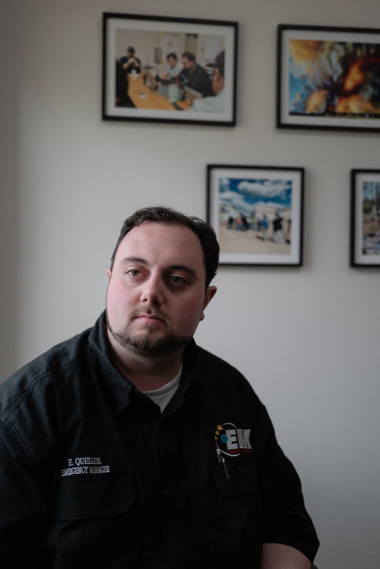 A man sits indoors in front of framed photographs on a white wall.