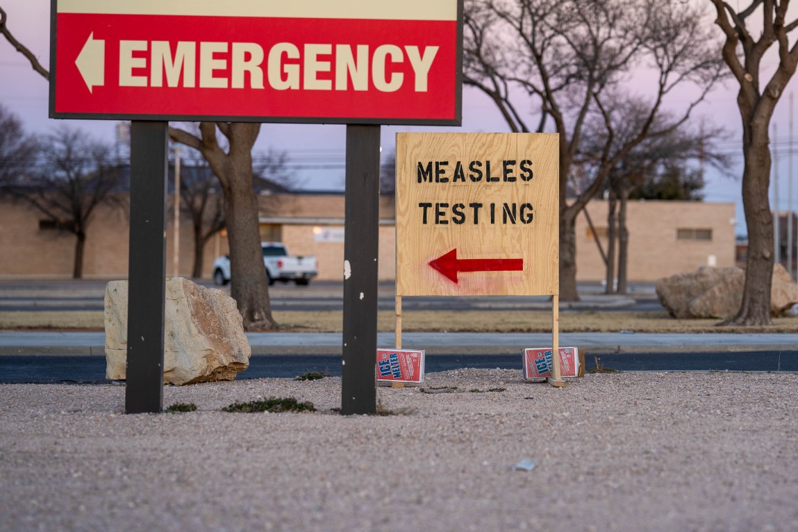 There is a large sign in the foreground pointing toward the direction of the emergency room of a hospital, while another sign in the background points to a measles testing location.