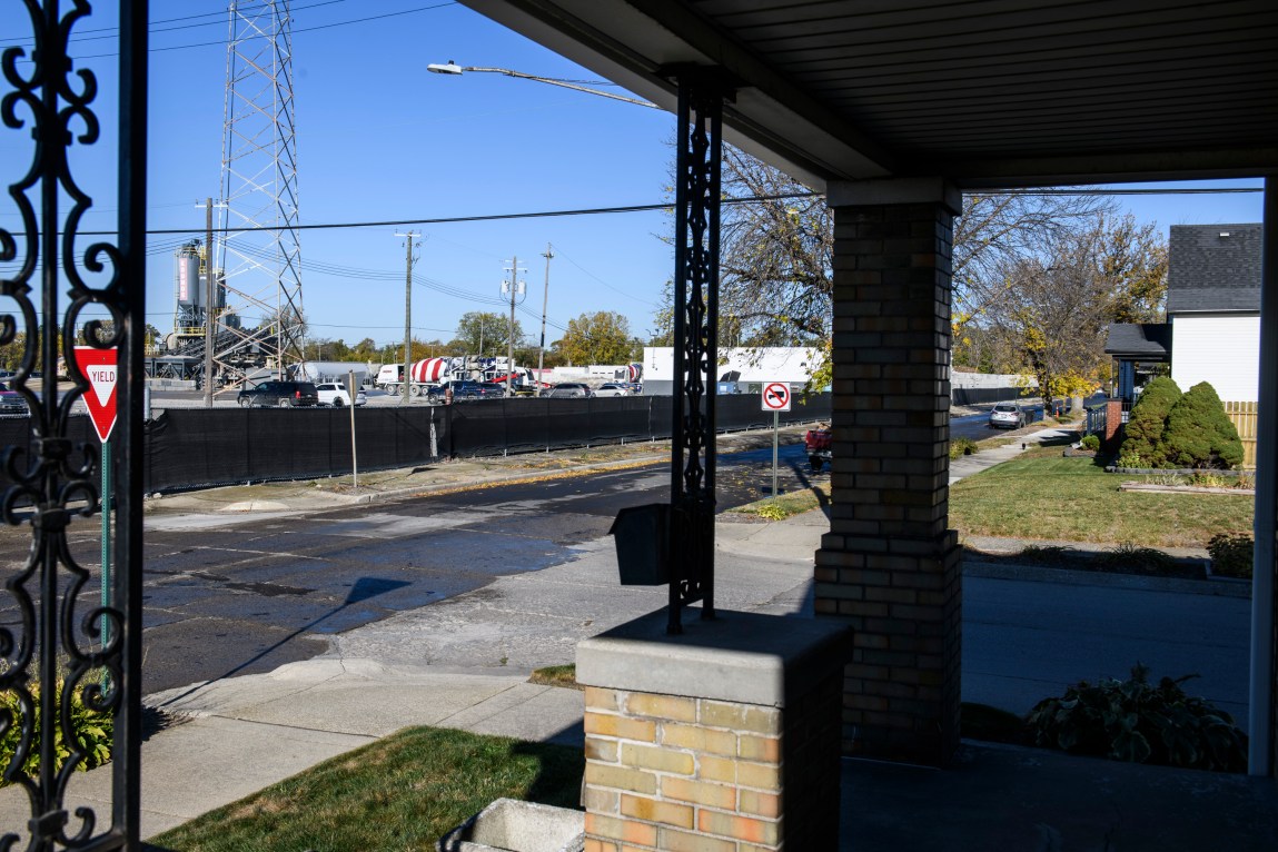 A view from a house front porch shows a street and a fenced-in lot with cars, concrete-mixing trucks and industrial buildings.