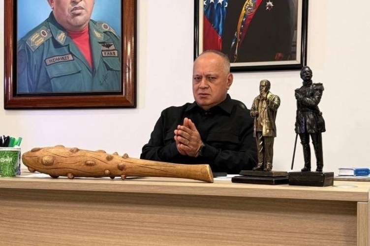 A man with a stern expression sits at a desk. A spiked club is on the desk in front of him.