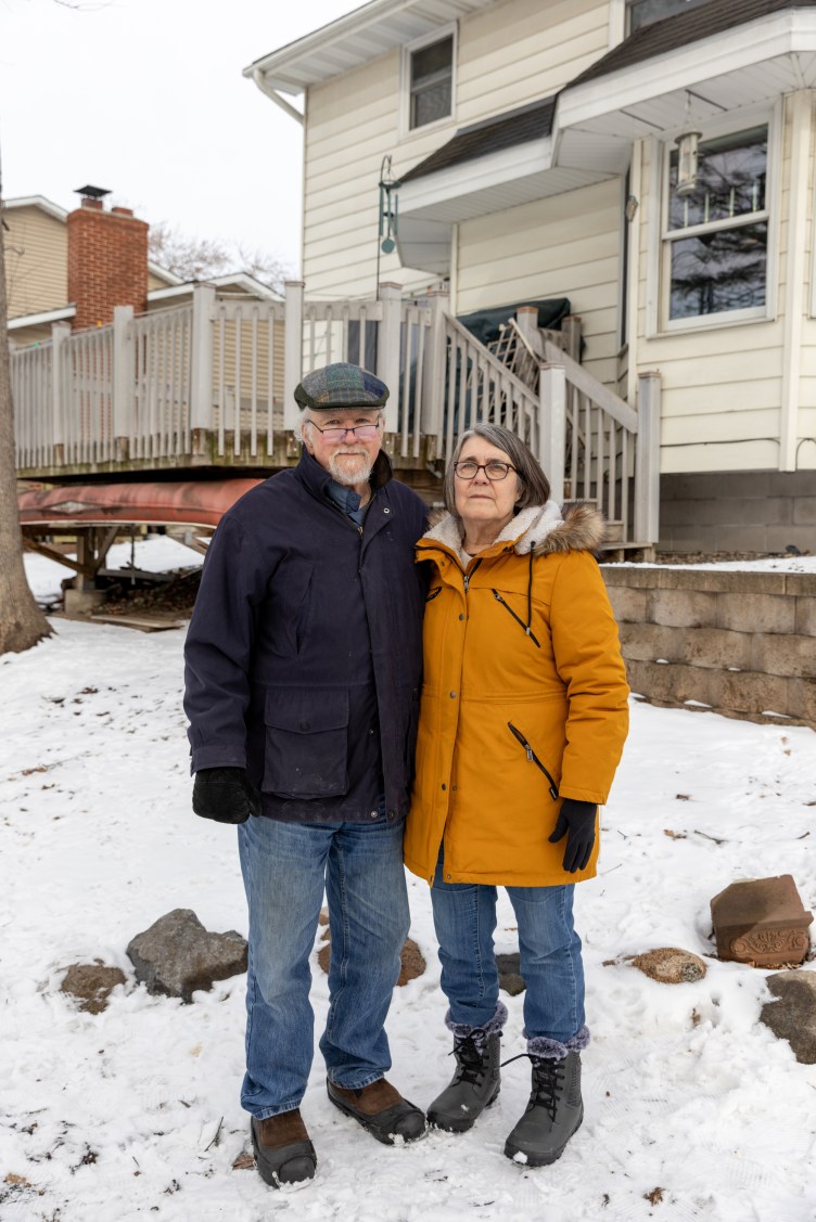 An elderly man and woman stand together outside in the snow, looking at the camera with stern expressions, with a back porch and a house in the background.