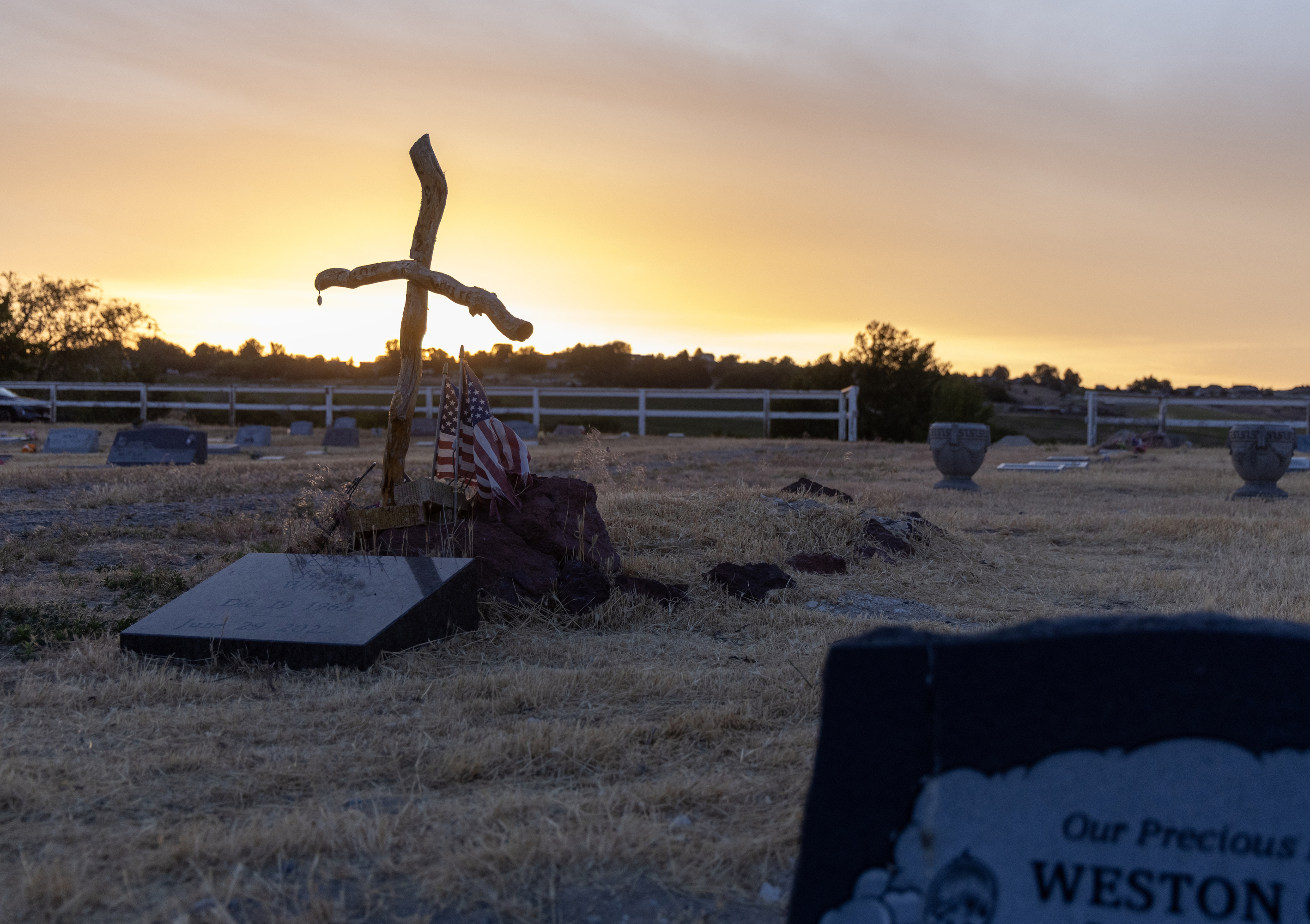 The sun rises behind a cemetery scattered with gravestones, a wooden cross and U.S. flags.