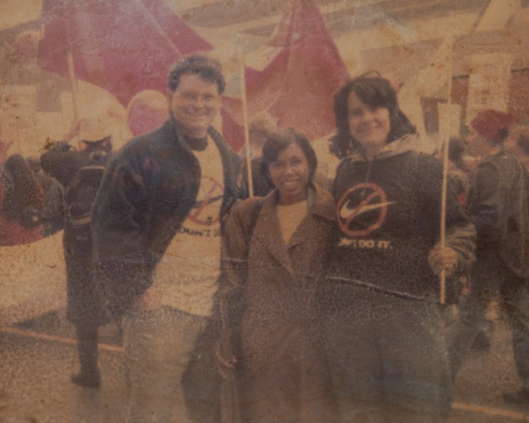 A faded and creased photograph showing a smiling, young Cicih standing between two smiling taller people wearing shirts with the Nike logo crossed out and the words “Don’t Do It.” Behind them is a march with people walking and holding banners and signs.