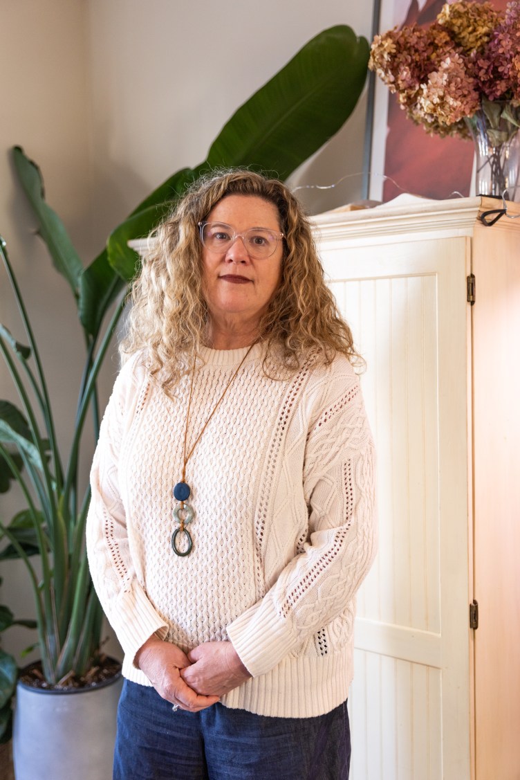 A woman stands looking at the camera with her hands clasped in front of her with a large houseplant behind her.