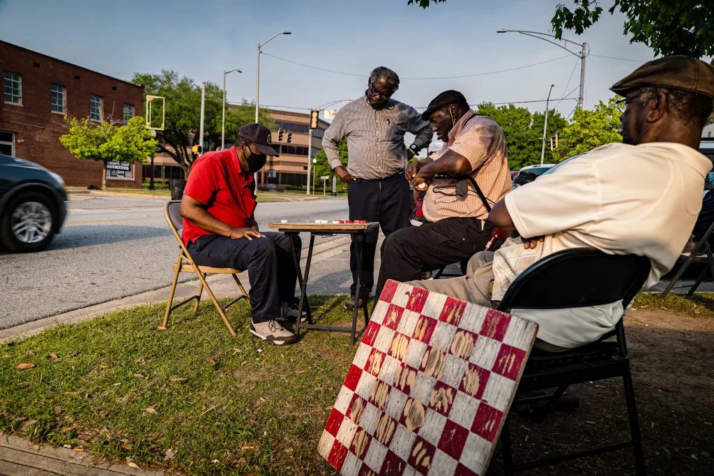 A group of men playing and observing a game of checkers. In the foreground, a worn wooden checkerboard leans against a man’s chair.