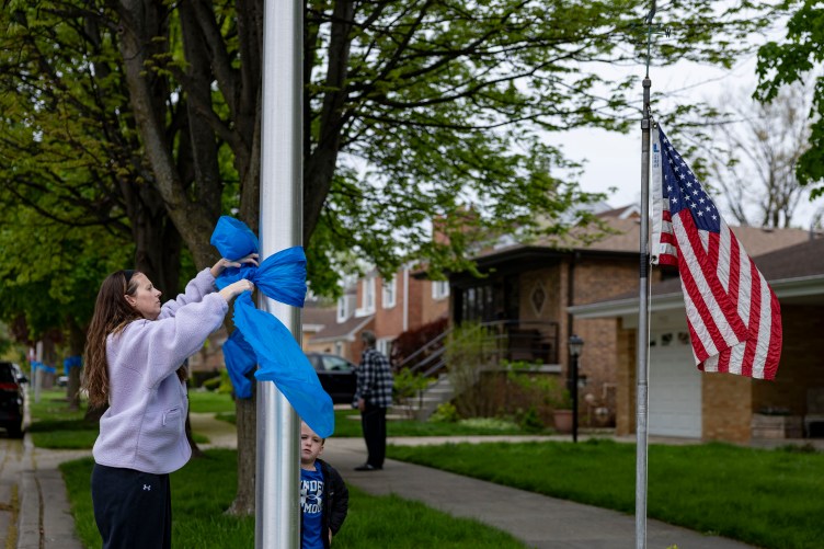 A woman in a lavender sweatshirt ties a blue ribbon around a metal pole in a residential neighborhood, while a young boy watches from nearby. To the right, an American flag hangs from a separate pole in front of a brick house.
