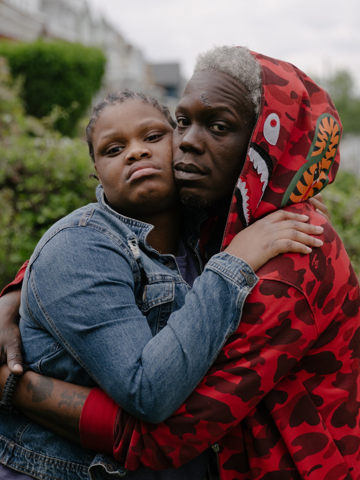 A daughter, wearing a jean jacket, and a father, wearing a brightly patterned red hoodie, pose holding each other and leaning their heads together.