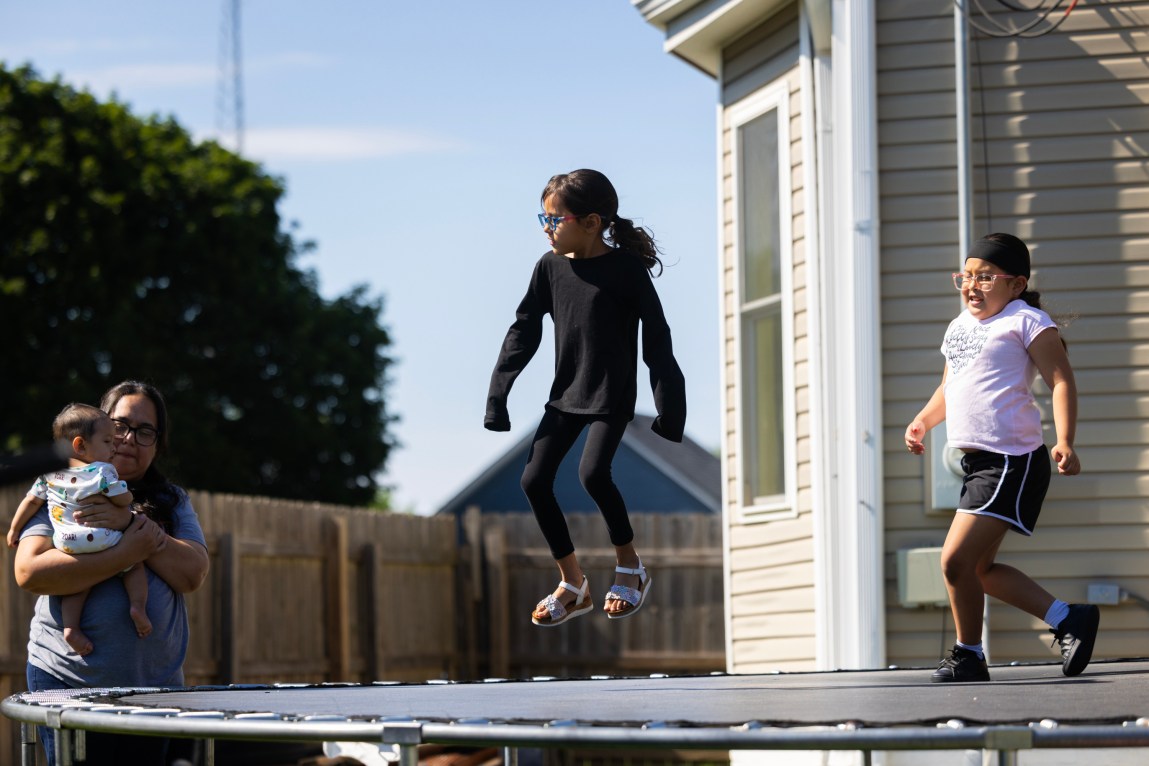 Two children jump on a trampoline outside. A woman stands nearby holding a baby. Behind the trampoline is a house with beige siding, a wooden fence and dark green vegetation.