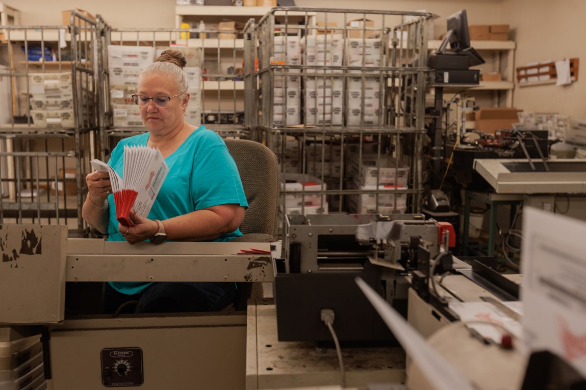 A woman wearing glasses and a teal T-shirt fans through a stack fo red-and-white ballot envelopes in an industrial workspace.