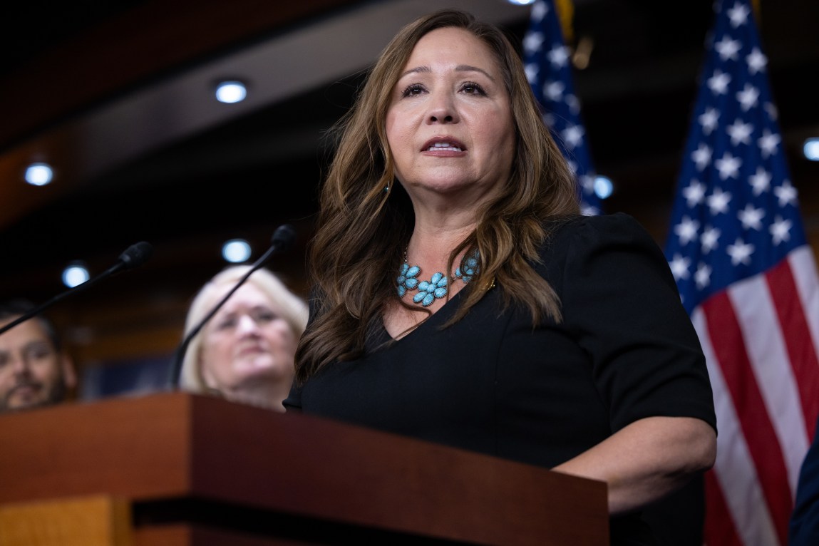A woman wearing a black dress and necklace made of turquoise speaks at a podium during a press conference. There are two out-of-focus onlookers and two United States flags in the background.