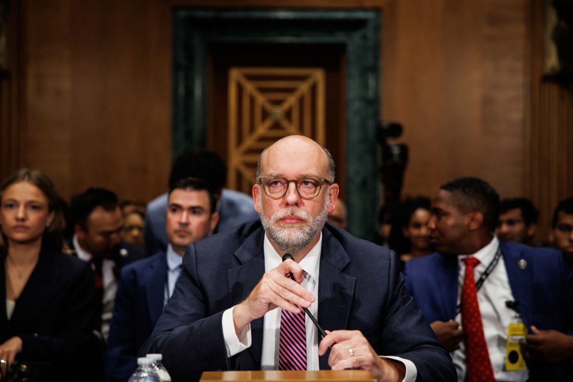 A man with a gray beard, glasses, suit and tie sits in front of a microphone in a hearing chamber.