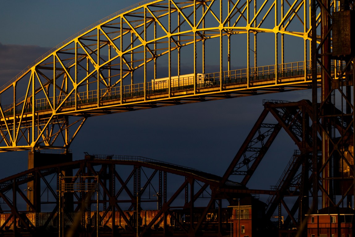 A white semitruck drives across a massive steel truss arch bridge that’s lit yellow by warm sunlight. Darker industrial bridge structures sit in shadow underneath against a deep twilight sky.