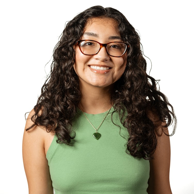 A woman with shoulder-length wavy dark brown hair smiles at the camera. She wears glasses, a green tank top and a silver necklace.