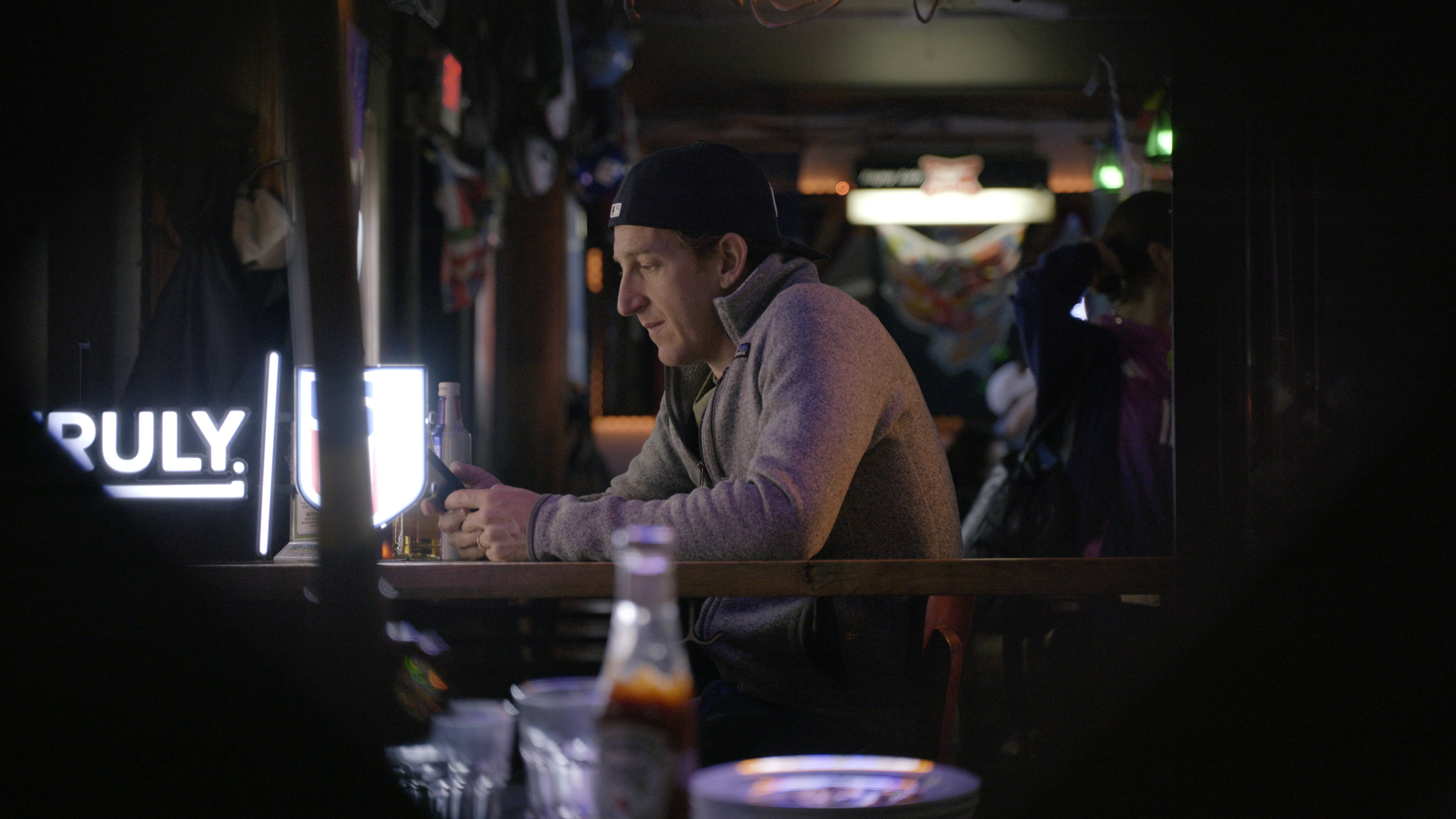 A man wearing a backward baseball cap sits in a dark bar, staring at his phone.