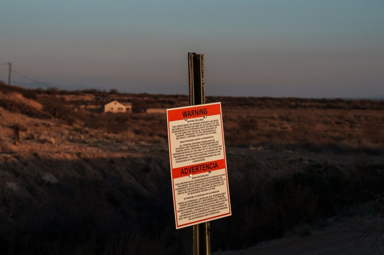 A sign on a post stating in English and Spanish that the area is a military zone, in front of a barren stretch of desert with a small white house.