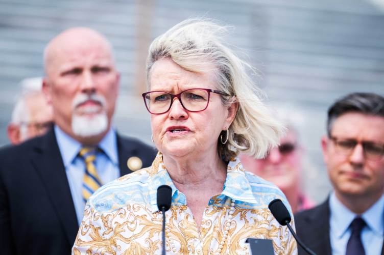 A woman wearing red glasses and a patterned blouse speaks into microphones at an outdoor press event with people gathered behind her.