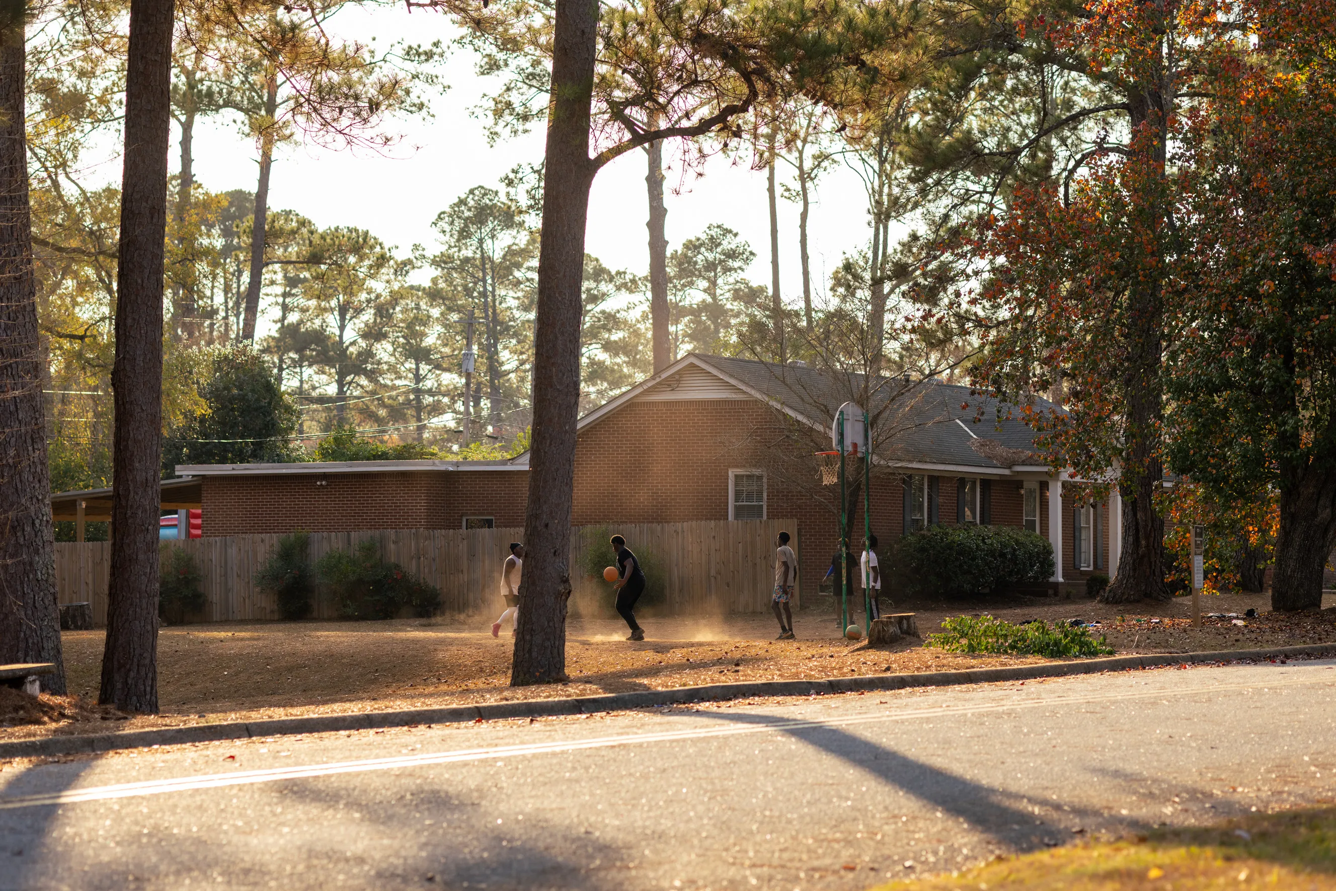 A group of boys plays basketball between trees and houses in a suburban neighborhood.