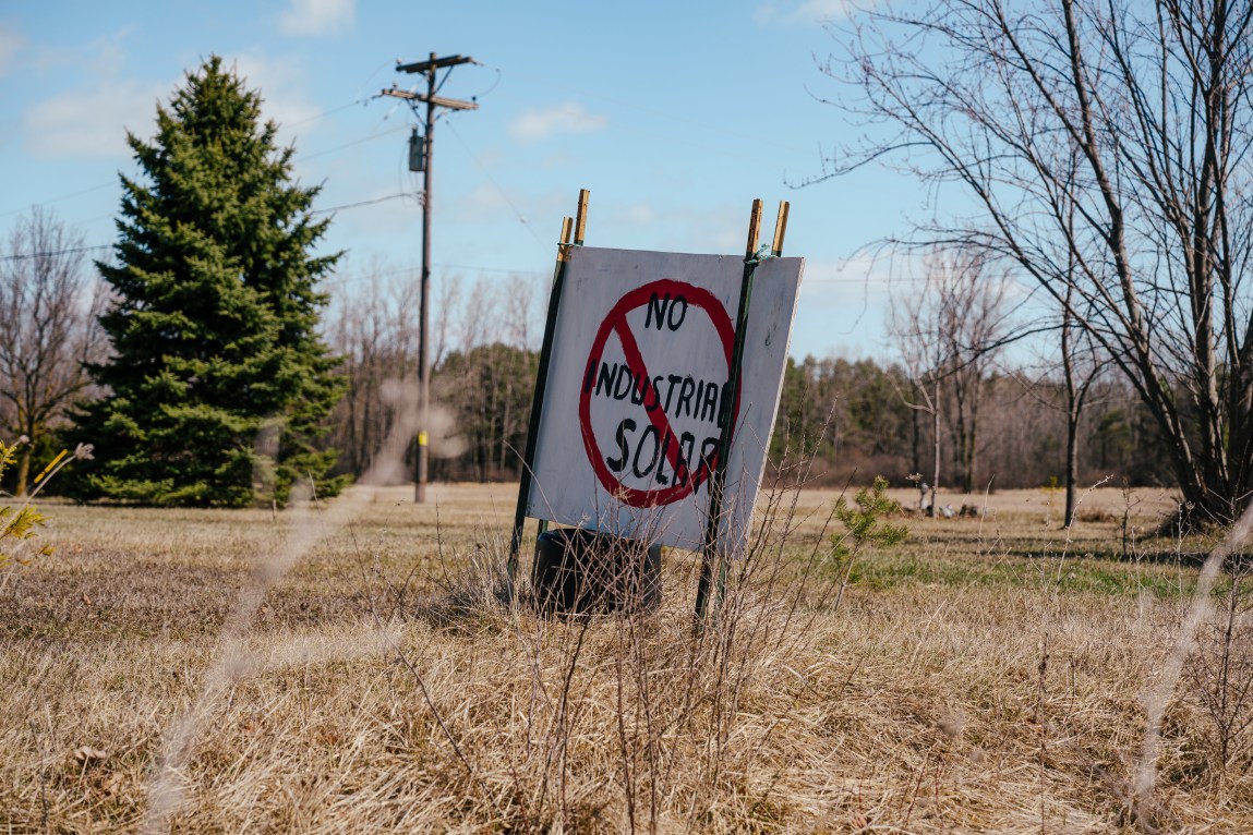 A wooden sign shows the hand-painted slogan “NO INDUSTRIAL SOLAR” circled in red with a line through it. The sign, held up by thin metal posts, is in a field with trees in the background.