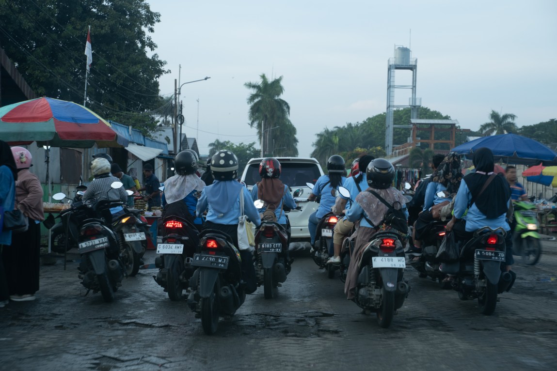A crowd of motorcycles on a potholed street. The road is flanked by stands under large beach umbrellas. Palm trees and other vegetation can be seen in the distance.