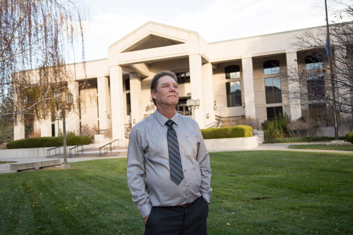 A man wearing a gray shirt, black pants and striped tie looks off into the distance in front of a green lawn and white building with pillars.