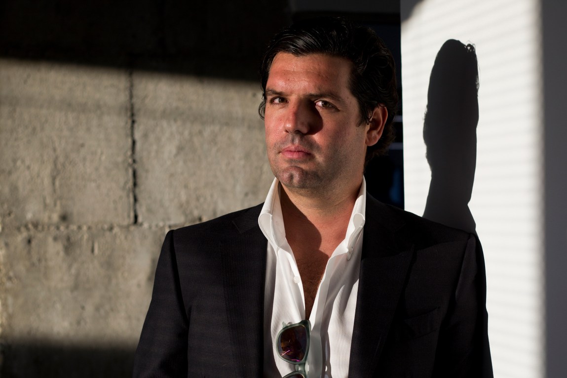 A man in a sportscoat looks at the camera. He is lit dramatically and casts a shadow on the wall behind him.