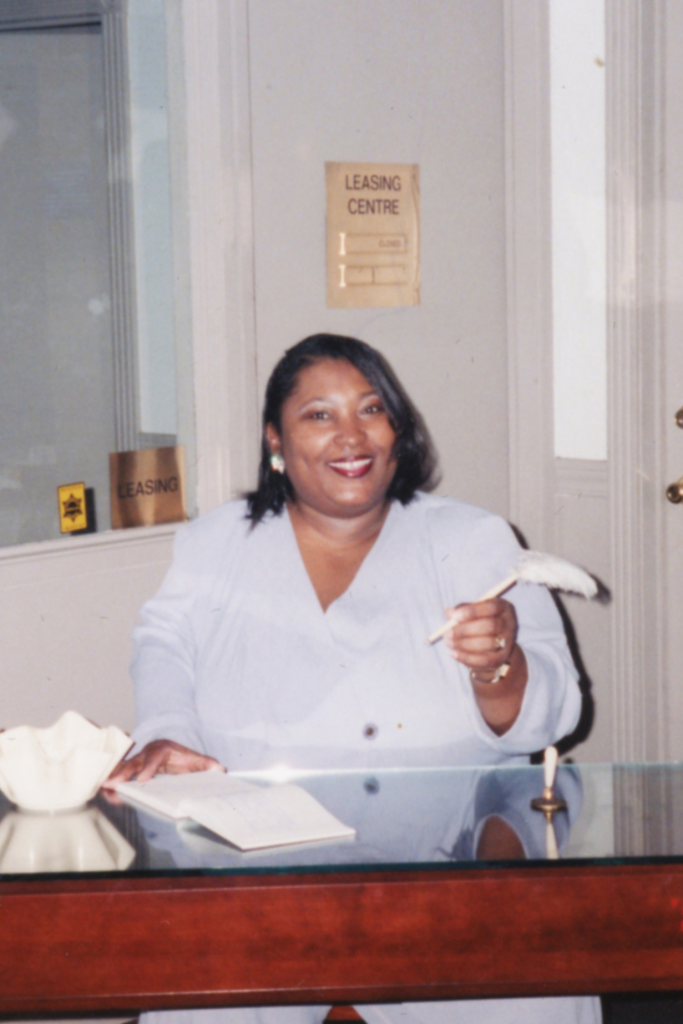 A woman is smiling and sitting at a wooden desk holding a pen.