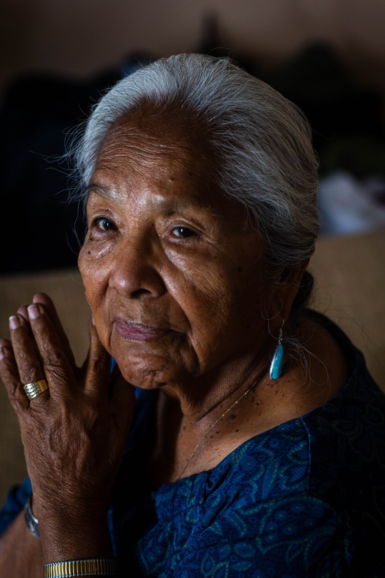 A close-up portrait of an elderly woman with silver hair tied back. She is wearing a blue patterned top, turquoise earrings and a gold ring, and holding her hands near her face.
