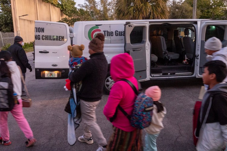 Children of varying ages walk toward the back of a large white van in the early morning.