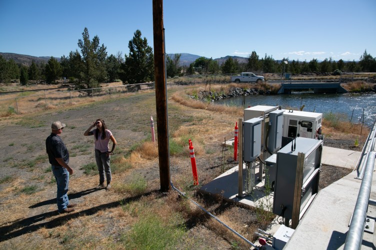 A man and a woman stand beside pipes and equipment next to a large body of water. 