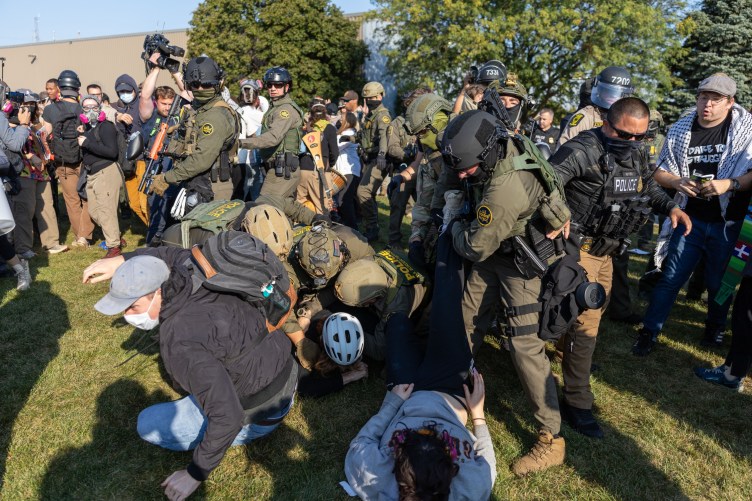 Federal immigration agents dressed in green combat gear and carrying crowd-control weapons confront a group of protesters. Some agents are shown tackling people to the ground.