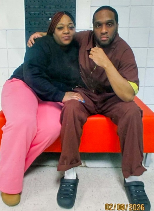 A man and woman pose for a photo while sitting on a red bench against a white tiled wall. The man is wearing a maroon jumpsuit and has his arm around the woman’s shoulder, while her hand is resting on his knee.