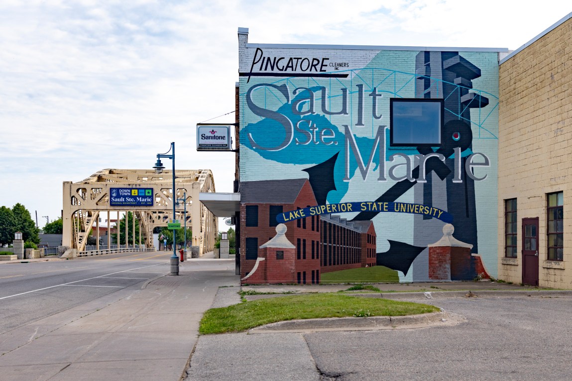 A street-level view shows a building adorned with a large Sault Ste. Marie mural that includes the phrases “Pingatore Cleaners Inc.” and “Lake Superior State University.” In the background, a light-yellow steel bridge spans a road under a partly cloudy sky.