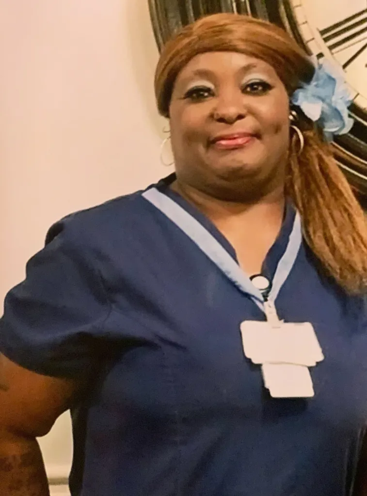 A woman is smiling with her scrubs on. She has blue eye shadow and a blue flower in her hair. 