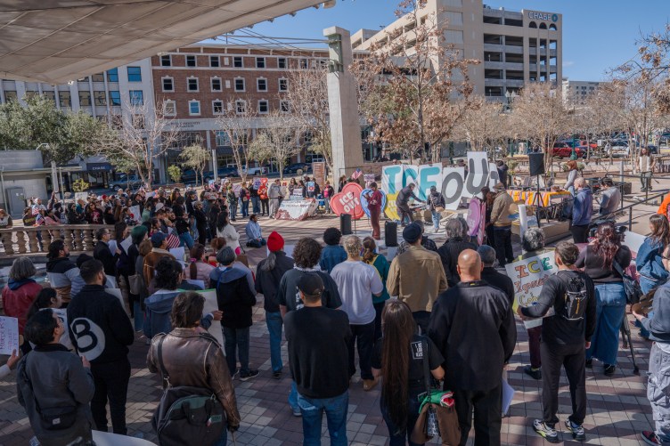 A large crowd of people gathers in an urban plaza for an outdoor demonstration. Activists hold large cutout letters spelling "ICE OUT" and carry signs in front of a speaker system, with surrounding city buildings visible in the background.