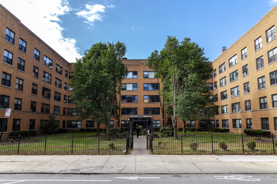 A large apartment building with a lawn and gate in front of it. There are two large trees framing the entrance of the building.