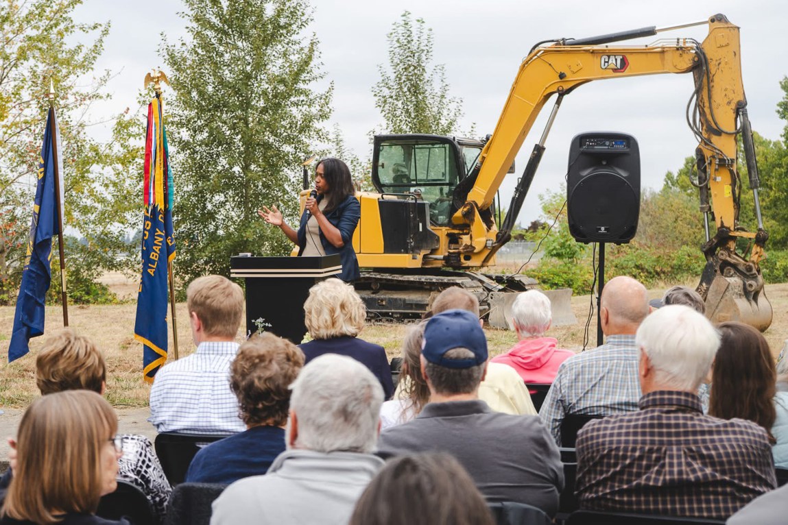 A woman speaks to a crowd at a lectern in front of an excavator.