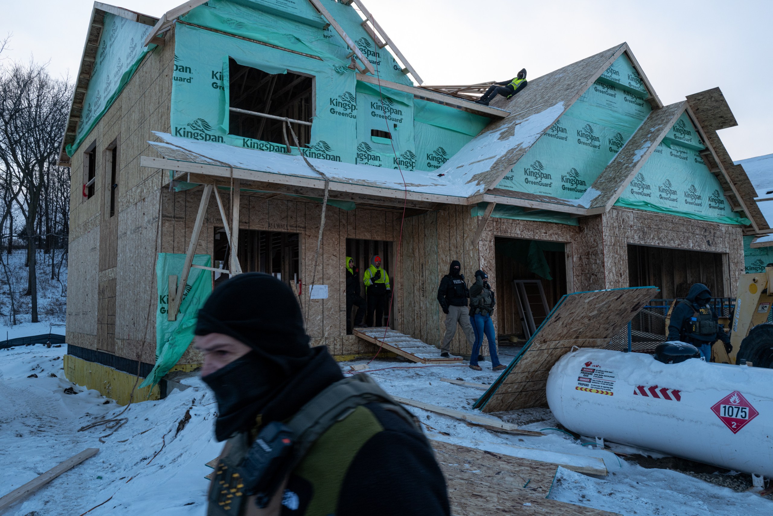 Men in tactical vests and ski masks walk out of the plywood shell of a house. A worker in a high-visibility vest leans against the peak of a roof as others on the ground watch the agents go.