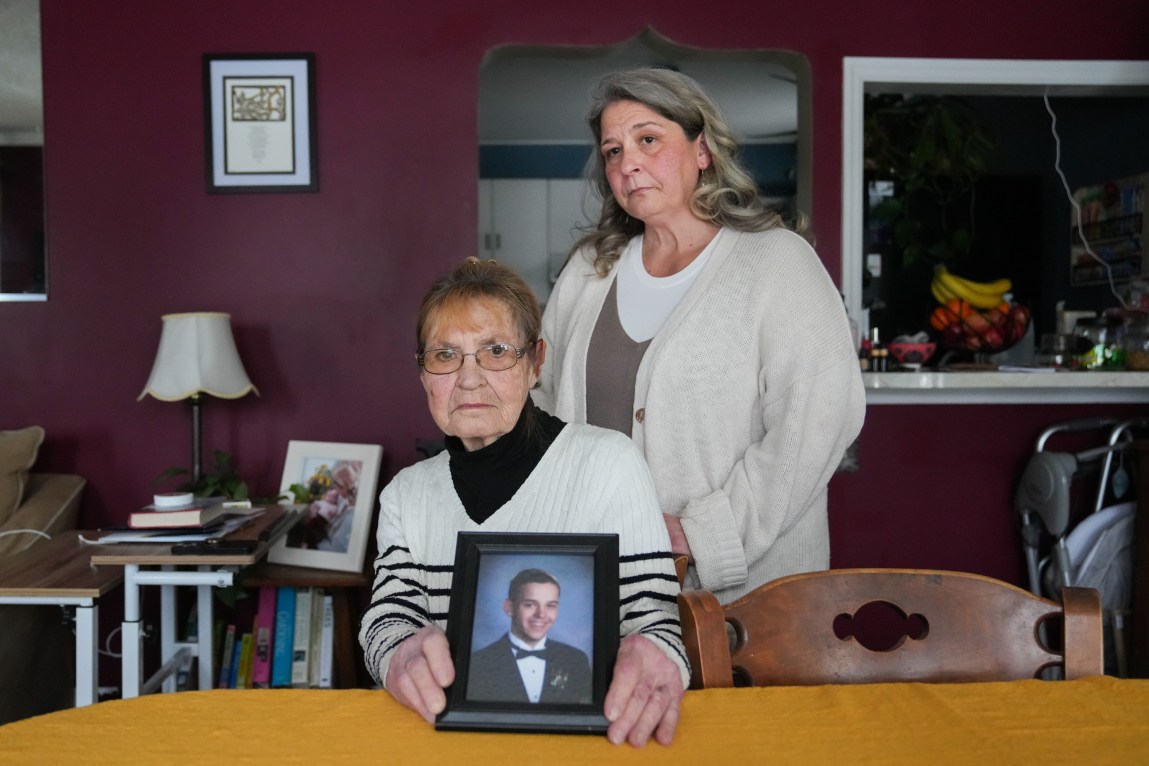A woman sits at a dining room table, looking at the camera with a serious expression, holding a framed photograph of a young man in a tuxedo. Another woman stands behind her.