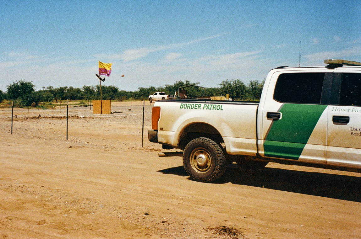 A white and green Border Patrol pickup truck drives on a dirt road past a campsite marked by a purple and yellow flag.