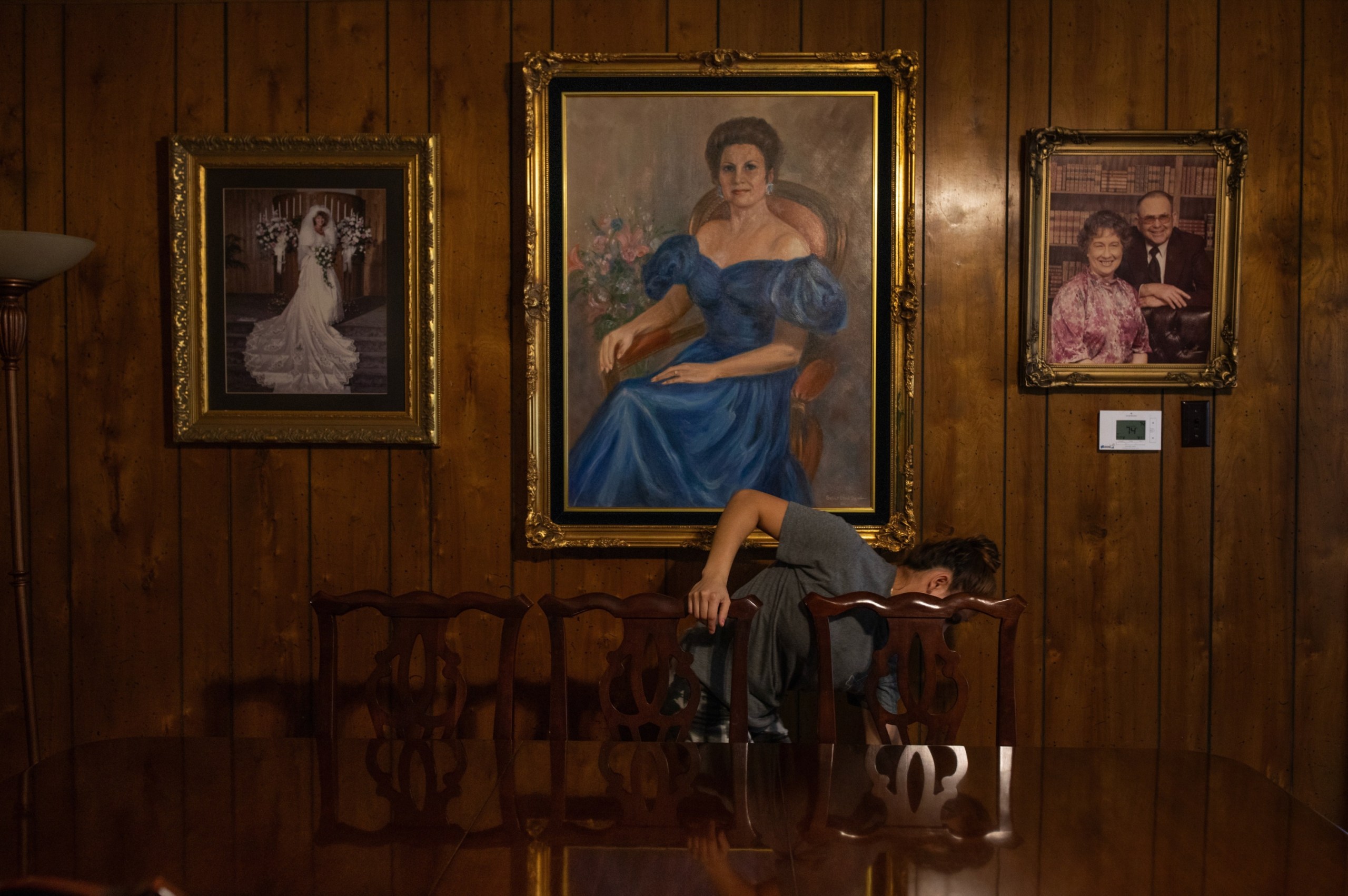A Latina woman bends down while cleaning a room paneled in dark wood, the back of a chair hiding her face, as portraits of well-dressed white people in elegant frames hang on the wall behind her.