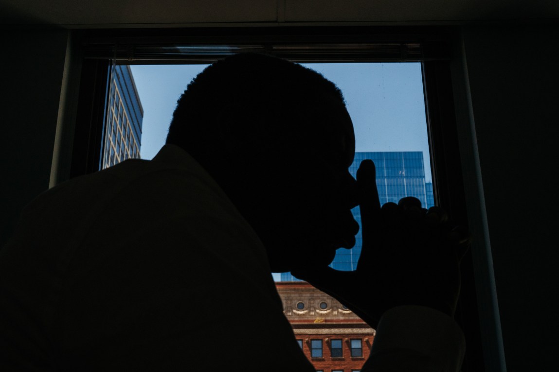 The silhouette of a man resting his chin and nose on his fingers, which are in an L-shaped position. The window behind him frames multiple skyscrapers.