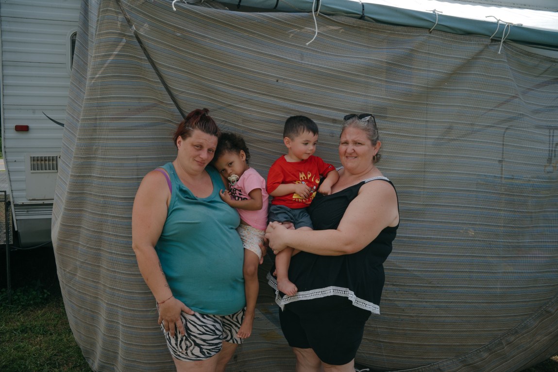 Two women stand in front of a cloth hanging in front of a camper. Both women hold children and look at the camera.