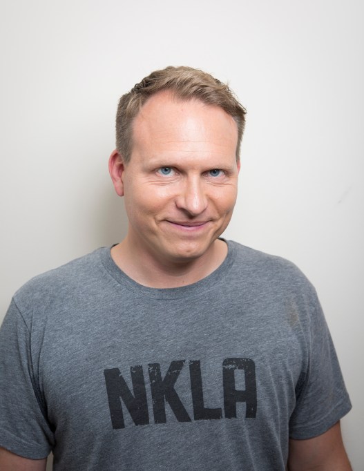 A man wearing a gray T-shirt smiles at the camera against a plain white background.