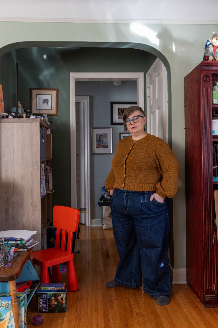 A woman stands with her hands in her pockets, looking at the camera, in the living room of her home. Next to her is a small table and small red chair for a child, with toys and books on the table and the floor. Behind her are family photos on the wall.