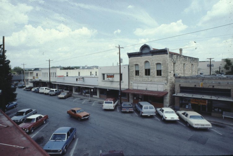 A high-angle view of a town street lined with storefronts and historic buildings, featuring numerous vintage automobiles parked along the road and curbs.