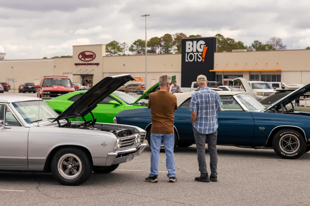 Two men standing in a strip-mall parking lot. On either side of them are two vintage cars with their hoods propped open.